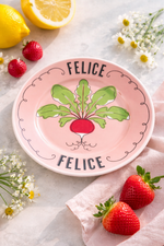 Piatto in porcellana rosa cipria Ø16 cm con scritta “Felice” e decoro ravanello, ambientato con fragole, limone e fiori su tavola elegante
