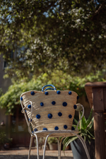 Borsa in paglia con pon pon blu appoggiata su sedia in ferro in giardino, stile naturale ed elegante.