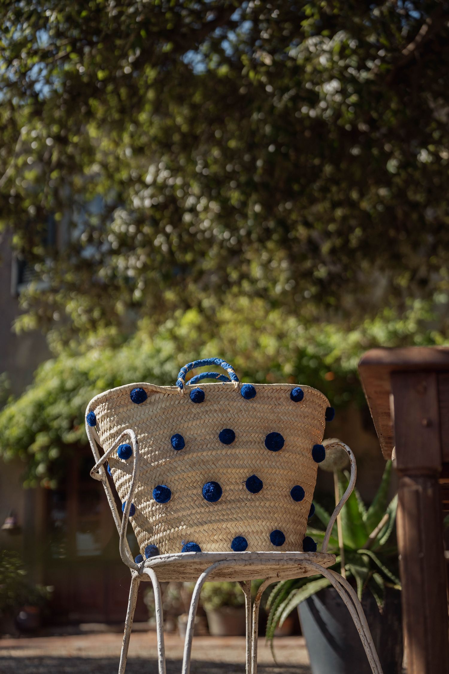 Borsa in paglia con pon pon blu appoggiata su sedia in ferro in giardino, stile naturale ed elegante.