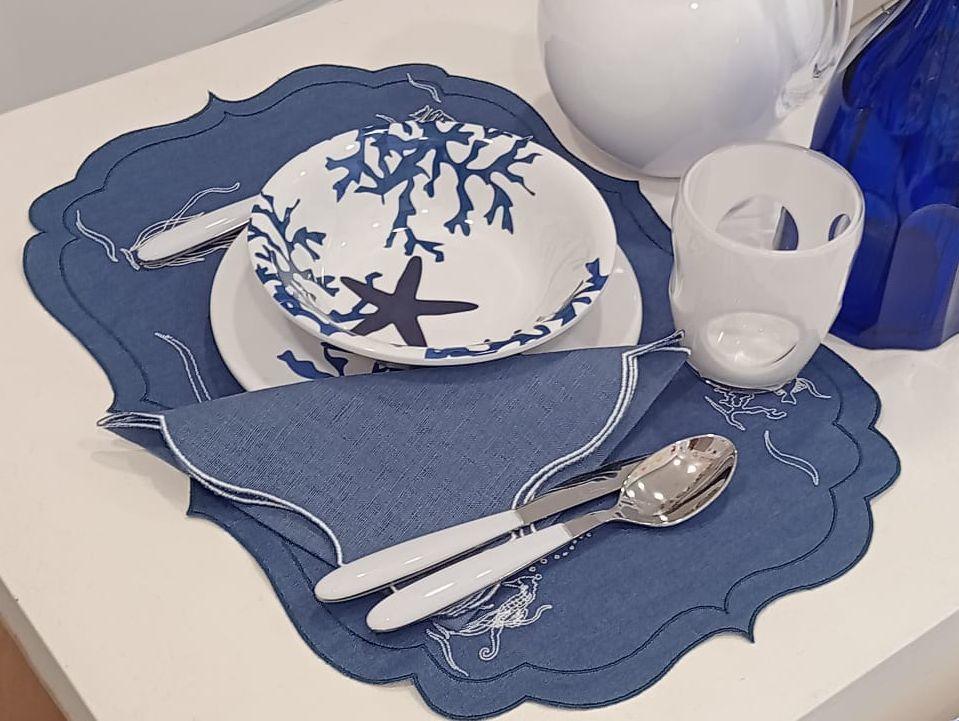 Mise en place estiva con americano e tovagliolo in lino cerato blu, ricami marini bianchi e piatti decorati con coralli, ideale per tavole eleganti al mare