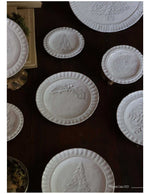 Collezione di piatti natalizi Virginia Casa in ceramica bianca con decori in rilievo di agrifoglio, pino e pigne, disposti su tavolo in legno in stile artigianale.
