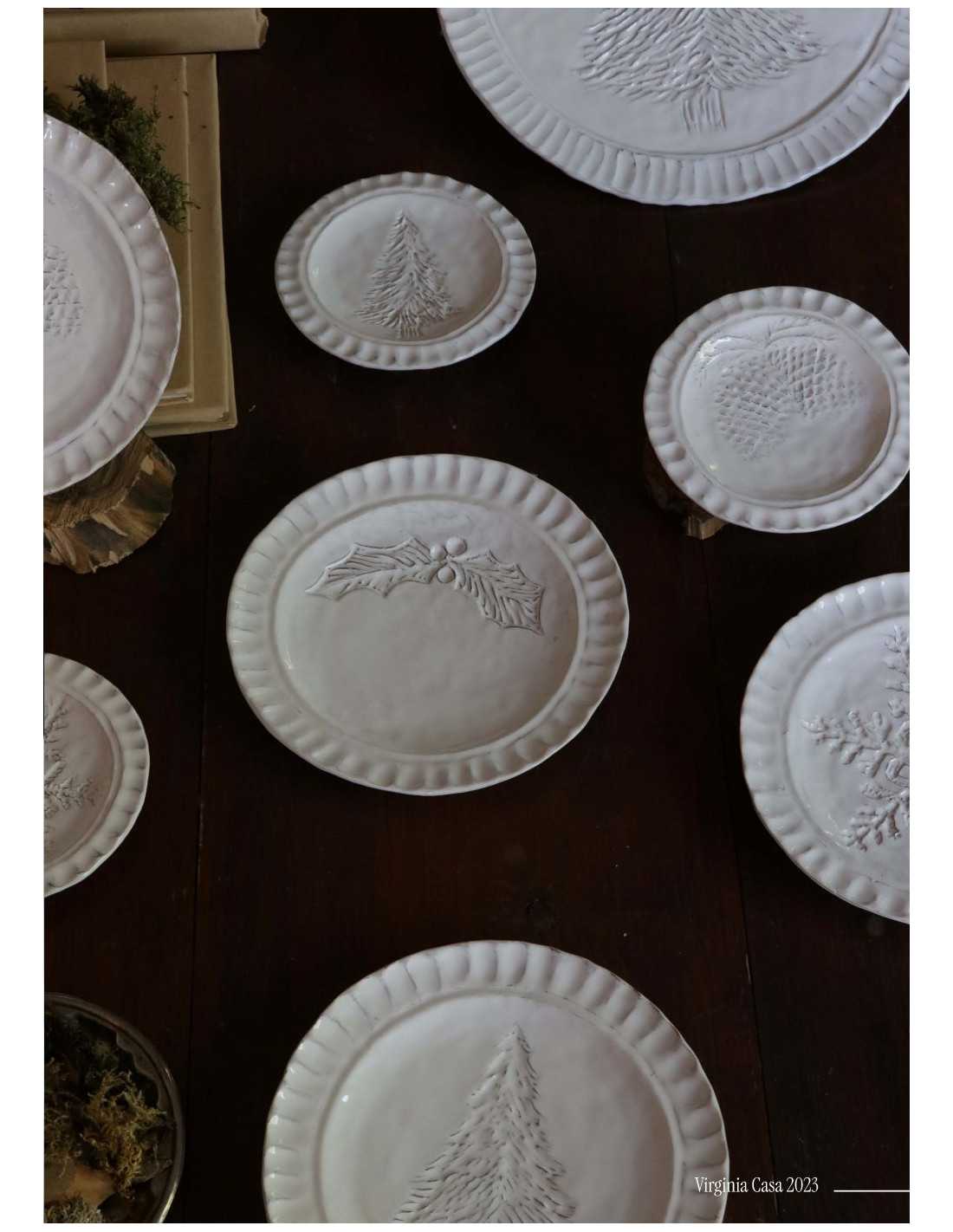 Collezione di piatti natalizi Virginia Casa in ceramica bianca con decori in rilievo di agrifoglio, pino e pigne, disposti su tavolo in legno in stile artigianale.