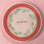 Piatto in porcellana Ø21 cm con scritta “piccole cose”, decoro foglie verdi e bordo rosso pesca su fondo rosa