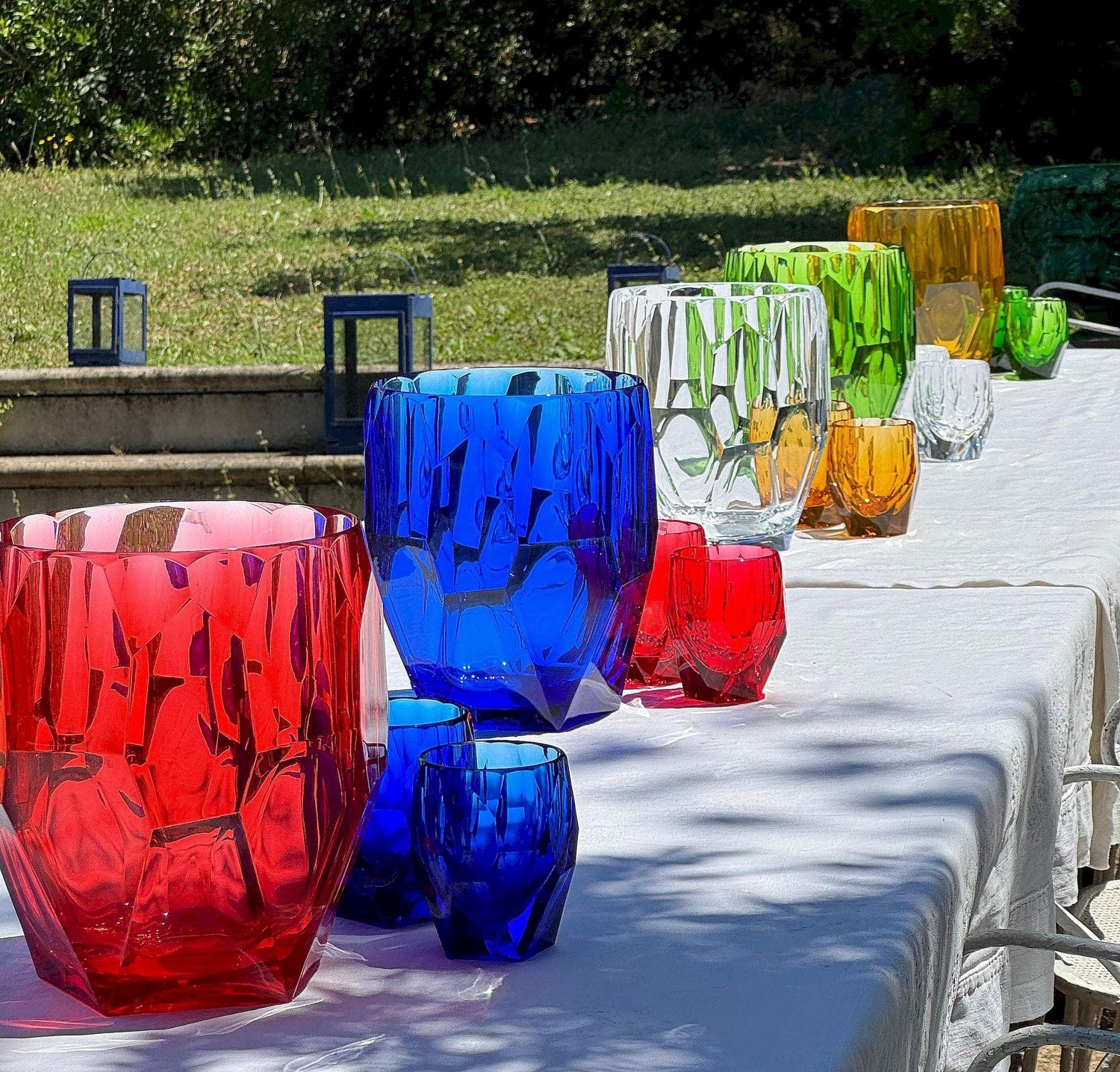 Porta bottiglie Antartica con bicchieri Milly e Super Milly di diversi colori di Mario Luca Giusti, in cristallo acrilico, fotografati all’aperto su tavolo estivo, design sfaccettato elegante e colorato
