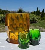 Porta bottiglie Antartica ambra con bicchieri Milly e Super Milly verdi di Mario Luca Giusti, in cristallo acrilico, fotografati all’aperto su tavolo estivo, design sfaccettato elegante e colorato