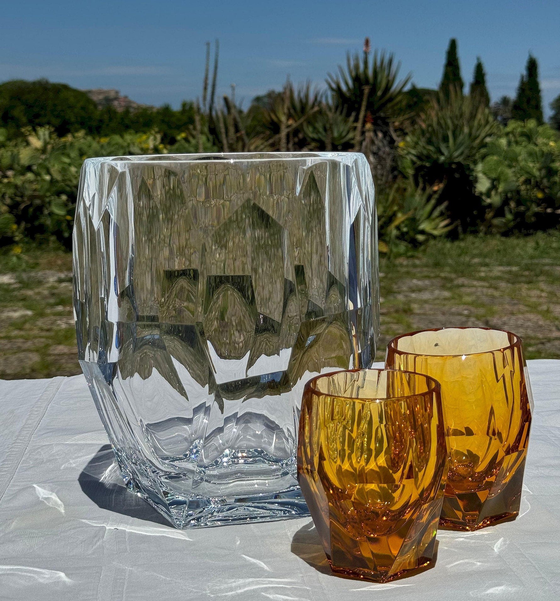 Porta bottiglie Antartica trasparente con bicchieri Milly e Super Milly ambra di Mario Luca Giusti, in cristallo acrilico, fotografati all’aperto su tavolo estivo, design sfaccettato elegante e colorato