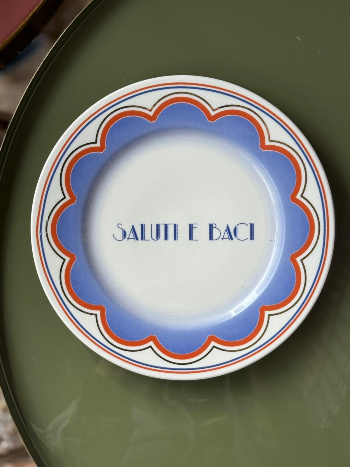 piatto Saluti e Baci diametro 21 cm in porcellana con decoro blu e rosso appoggiato su tavolo verde