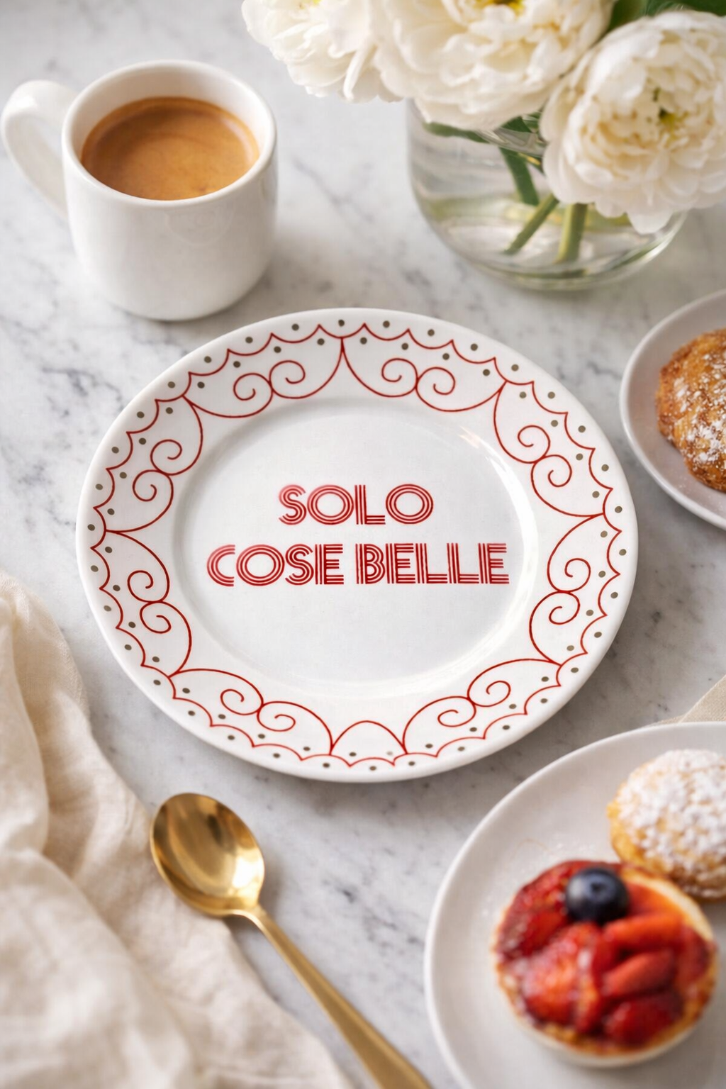 Piatto in porcellana bianco Ø16 cm con scritta “Solo Cose Belle” e decoro rosso ornamentale, apparecchiato su tavolo in marmo con caffè espresso, fiori bianchi, dolci e cucchiaino dorato.