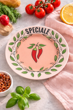 Piatto in porcellana rosa cipria Ø16 cm con scritta “Speciale” e decoro peperoncini rossi, ambientato con pomodori, basilico e limone su tavola elegante