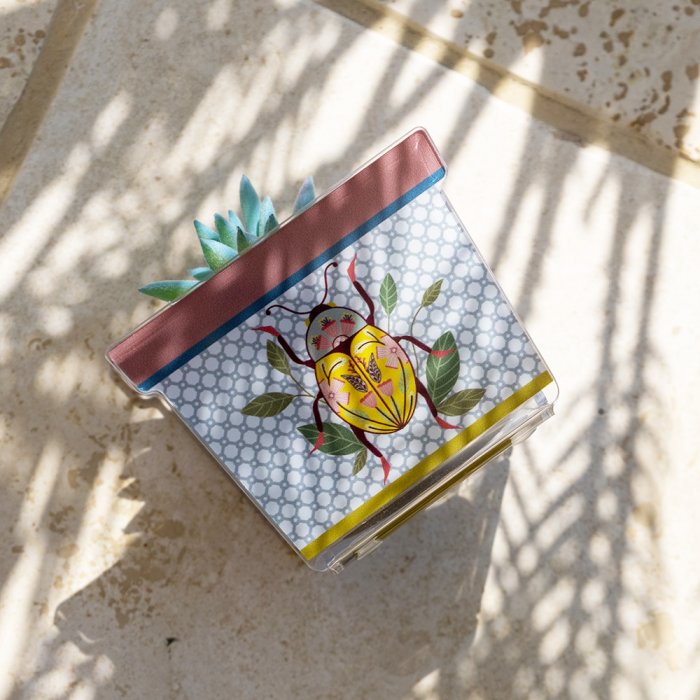 Vasetto JOY in acrilico decorato con scarabeo giallo e foglie verdi, fotografato dall’alto in luce naturale con ombre morbide.