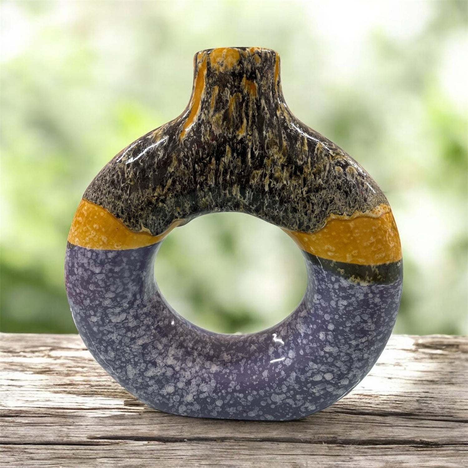 Vaso decorativo in ceramica a forma di anello con finitura lucida, colori viola, giallo ocra e nero marmorizzato, esposto su superficie in legno.