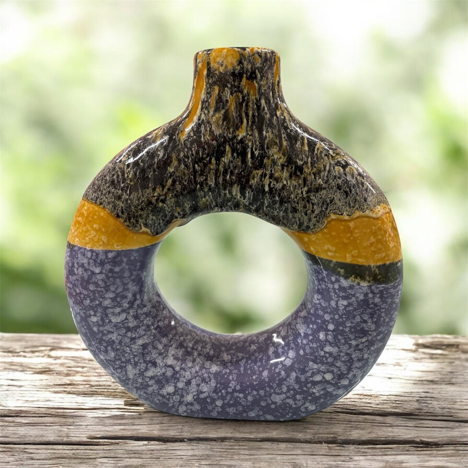 Vaso decorativo in ceramica a forma di anello con finitura lucida, colori viola, giallo ocra e nero marmorizzato, esposto su superficie in legno.