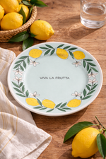 piatto Viva la Frutta limoni diametro 21 cm ambientato su tavolo in legno con limoni freschi e tovaglietta mediterranea